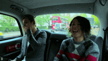 two men are sitting in a car with a red double decker bus behind them