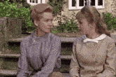 two women are sitting on a bench talking to each other in front of a building .