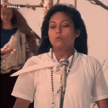 a woman is singing into a microphone while wearing a white shirt and necklace .