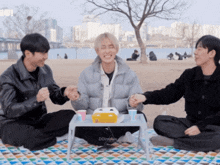 three young men are sitting on a blanket with a small table and a box that says dd