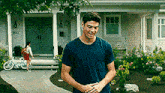 a man in a blue t-shirt is standing in front of a house with his hands on his hips .