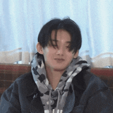 a young man wearing a black jacket and a camo hoodie is sitting on a couch