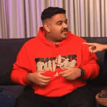 a man in a red hoodie is sitting on a couch talking to someone .