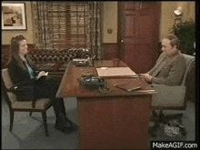 a man and a woman are sitting at a desk in an office talking to each other .