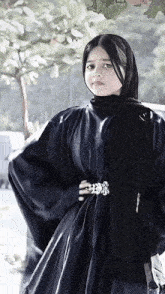 a woman wearing a black abaya and a black scarf is standing in front of a tree
