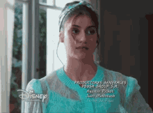 a woman in a blue dress is standing in front of a window with disney channel written on the bottom of the screen .