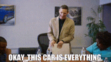 a man in a suit stands in front of a table and says okay this car is everything .