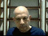 a bald man with a beard wearing a blue shirt looks at the camera