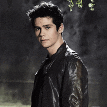 a young man in a black leather jacket stands in front of a dark wall
