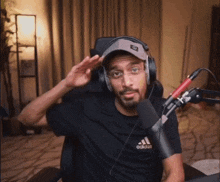 a man wearing headphones and a black adidas shirt salutes in front of a microphone