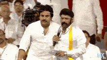 a man with a mustache is holding a microphone while standing next to another man with a yellow sash