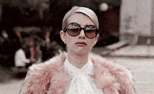 a woman wearing sunglasses and a pink fur coat looks at the camera