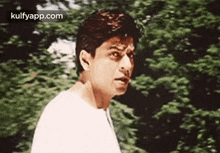 a man in a white shirt is standing in front of a tree and making a funny face .