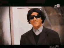 a man wearing sunglasses and a suit is standing in front of a 2m logo