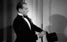 a black and white photo of a man in a tuxedo holding a hat .