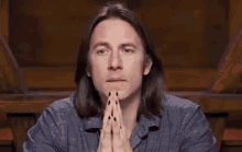a man with long hair and black nails is sitting at a table with his hands folded in prayer .