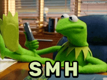 kermit the frog is sitting at a desk holding a cell phone and the word smh is above him