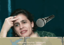 a woman holds her hand to her forehead in front of a microphone that says ' mestre sophia tomozelli '