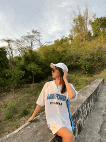 a woman wearing an ashford pharma shirt is sitting on a stone wall