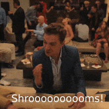a man in a suit is sitting at a table in front of a crowd with the words shroooooooms written on the bottom right