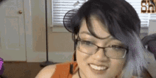 a woman with glasses and purple hair is smiling in front of a window .