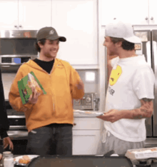 a man wearing a white shirt with a smiley face on it is talking to another man in a kitchen