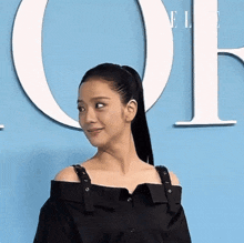 a woman wearing a black off the shoulder top stands in front of a blue wall with the letter o on it