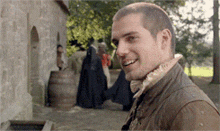 a man in a brown jacket is smiling in front of a stone wall