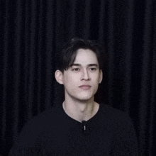 a young man in a black shirt is standing in front of a black curtain and waving his hand .