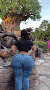 a woman in a black top and blue jeans is standing in front of a tree