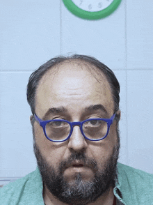a man with glasses and a beard looks at the camera in front of a green clock