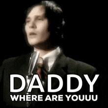 a man in a suit and tie is singing into a microphone with the words `` daddy where are youu '' behind him .