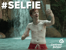 a shirtless man taking a selfie in front of a waterfall with #selfie written above him