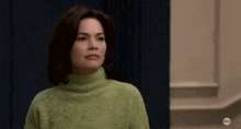 a woman in a green sweater is standing in front of a blue curtain and looking at the camera .