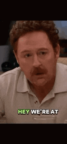 a man with a mustache is sitting at a table with a glass of water and saying hey we 're at .