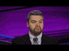 a man with a beard wearing a suit and tie looks to his left