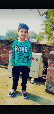 a young boy wearing a shirt that says roar stands in front of an lg air conditioner