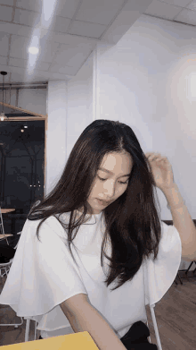 a woman in a white top sits at a table with her hand in her hair