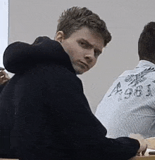 a man in a black hoodie is sitting at a desk in a classroom with other people .