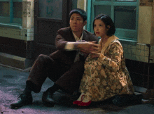 a man and a woman sit on the sidewalk in front of a building with a sign that says ' a '