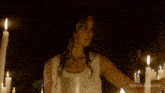 a woman in a white tank top stands in front of a row of lit candles with nemo-beatrice written in the corner