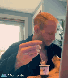 a man is eating yogurt with a spoon while a dog looks on in front of a momento logo