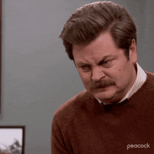a man with a mustache is wearing a brown sweater with peacock written on it .
