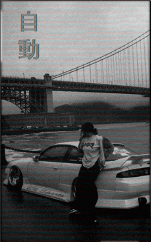 a black and white photo of a man leaning on a car with a bridge in the background