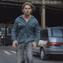 a man in a blue shirt is walking down a street with #caughtstealingmovie on the bottom