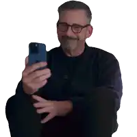 a man with glasses and a beard is sitting down looking at his phone