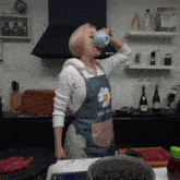 a woman wearing a blue apron with a flower on it drinks from a cup