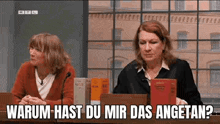 two women are sitting at a table in front of a microphone with a sign that says warum hast du mir das angetan ?