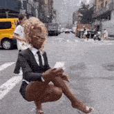 a woman in a suit and tie is squatting on the street looking at her phone