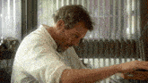 a man in a white shirt is sitting in front of a window with vertical blinds